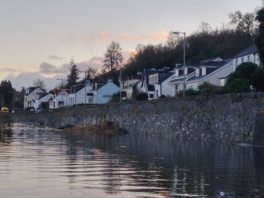 A waterfront property with a stone wall, white houses, and a calm body of water reflecting the sky.