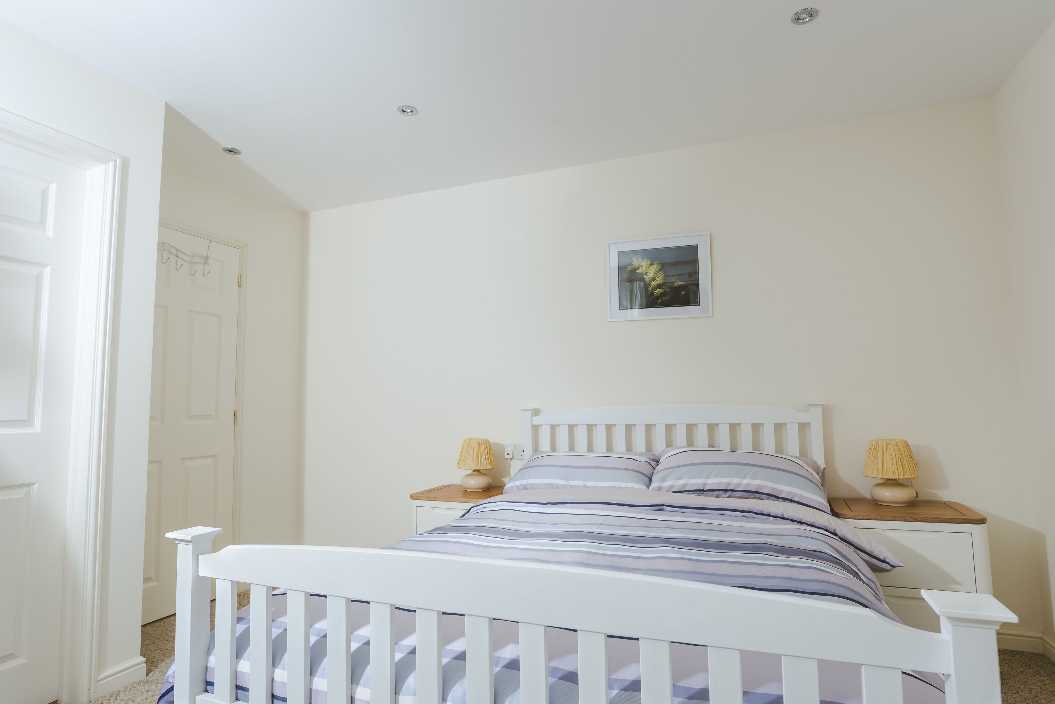 A white bed with a striped bedspread and two bedside lamps, a small picture on the wall, and a white door in the background.