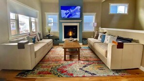 Una sala de estar con dos sofás blancos, una mesa de centro de madera, una chimenea y un televisor montado sobre ella.