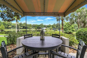 A covered outdoor seating area with a table and chairs, overlooking a lake and park.