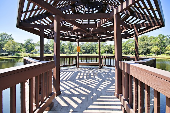 Ein Pavillon mit Holzboden und Geländern, umgeben von Wasser und Grünflächen.