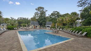 A rectangular swimming pool with a tiled deck, surrounded by lounge chairs and small buildings with sloped roofs. - Sunset Rentals | Evian 170 (Hilton Head Island)