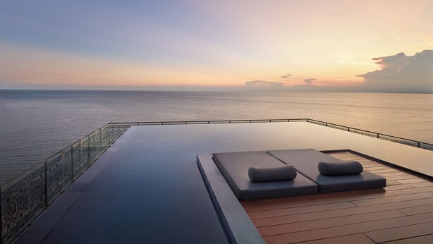 An infinity pool with lounge chairs and a glass railing overlooking the sea at sunset. - Skool Plus Vung Tau (Ho Chi Minh City)