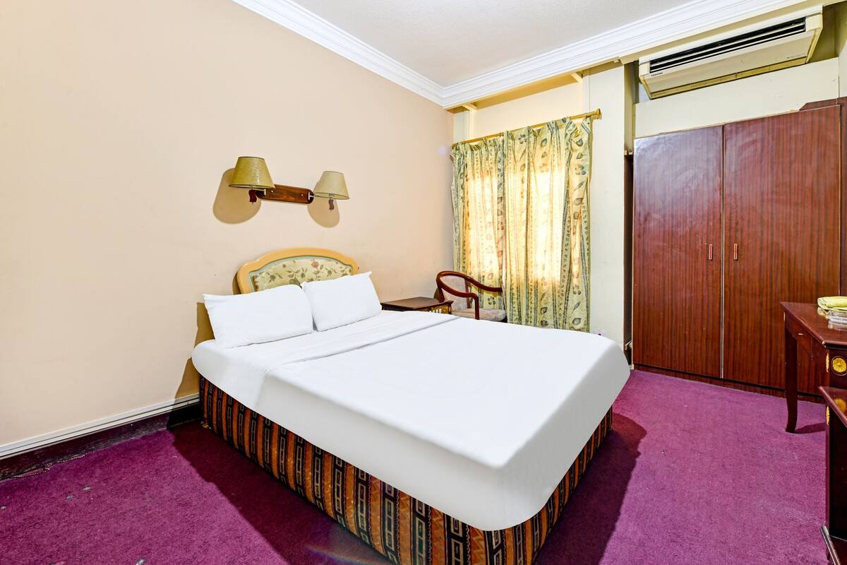 A hotel room with a large bed, a wooden wardrobe, a desk with a lamp, and a chair.