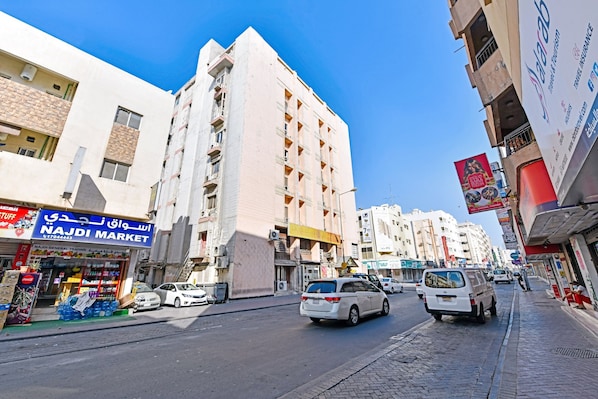 A street view with a white van parked on the side, a building with a sign reading "NAJDI MARKET", and another building with a sign for "Travel Insurance".