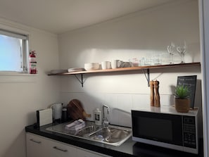 Una cocina con un lavamanos, microondas, estante con vajilla y vasos, extintor y una pizarra con un recordatorio.