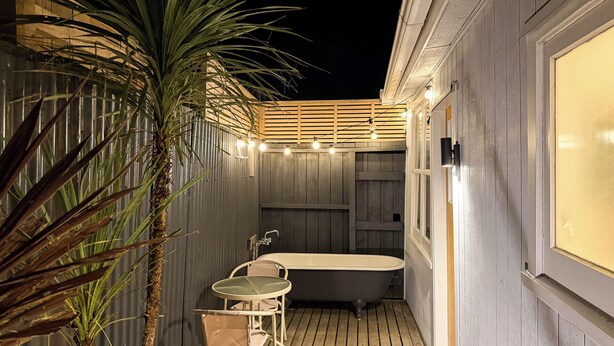 Une petite salle de bain extérieure, à l’abri, avec une baignoire, une petite table et des chaises, le tout entouré de murs en bois et d’un palmier.