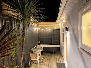 A small, enclosed outdoor bathroom with a bathtub, a small table, and chairs, surrounded by wooden walls and a palm tree.