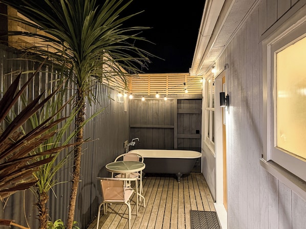 Une petite salle de bain extérieure, à l’abri, avec une baignoire, une petite table et des chaises, le tout entouré de murs en bois et d’un palmier.