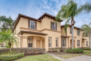 A two-story building with a beige facade, multiple windows with shutters, and a landscaped front yard with palm trees and shrubs.