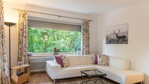 Sala de estar com sofá branco, mesa de centro de vidro, radiador, janela com cortinas e fotografia de veado fixada na parede.