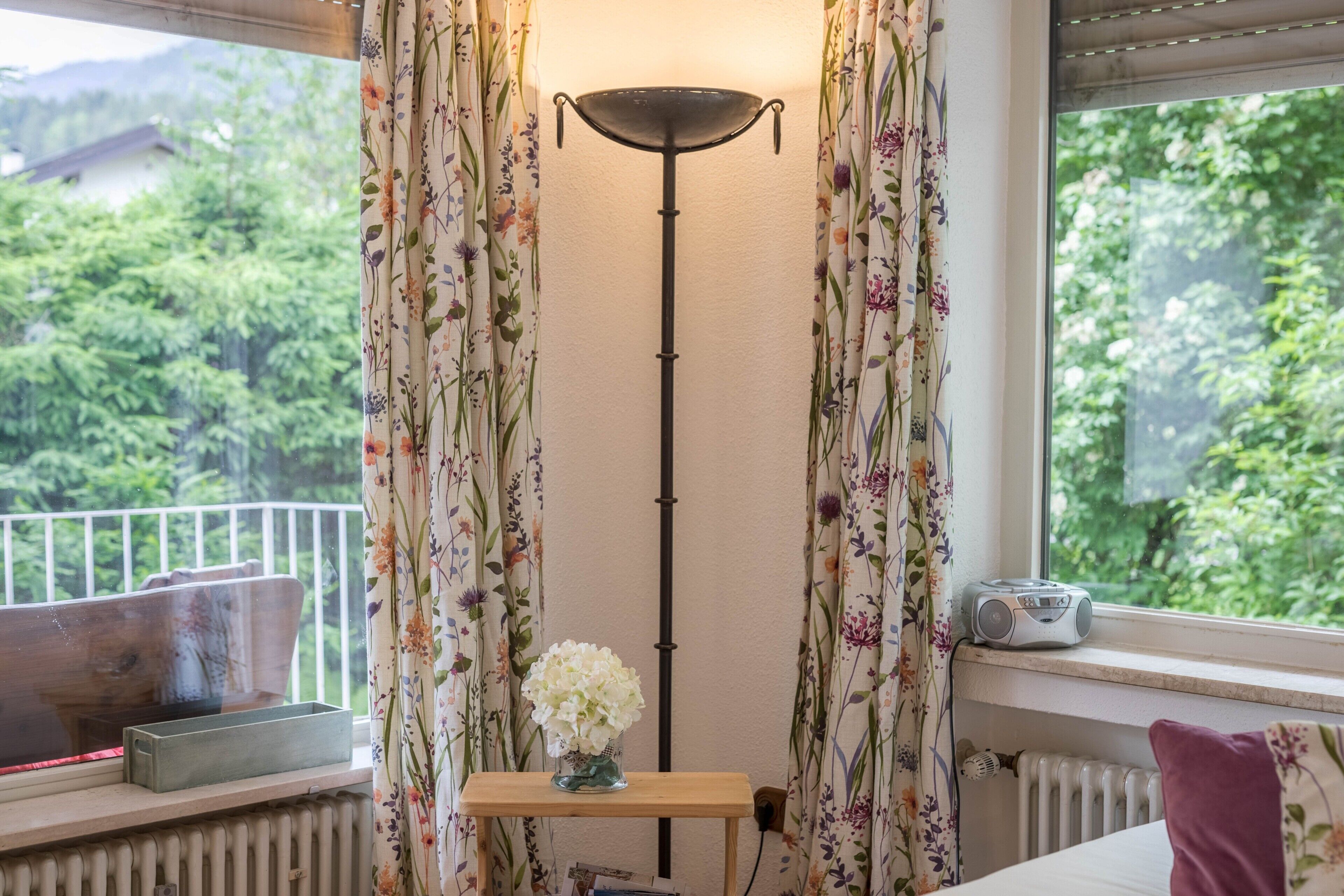 Une pièce avec un radiateur, une table en bois sur laquelle est posé un vase de fleurs, une radio et des rideaux à motifs floraux.