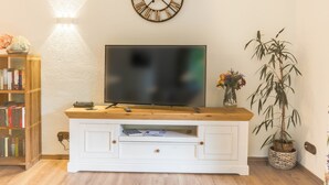 Uma sala de estar com televisão sobre um suporte de madeira, uma estante, um relógio e um vaso com planta.