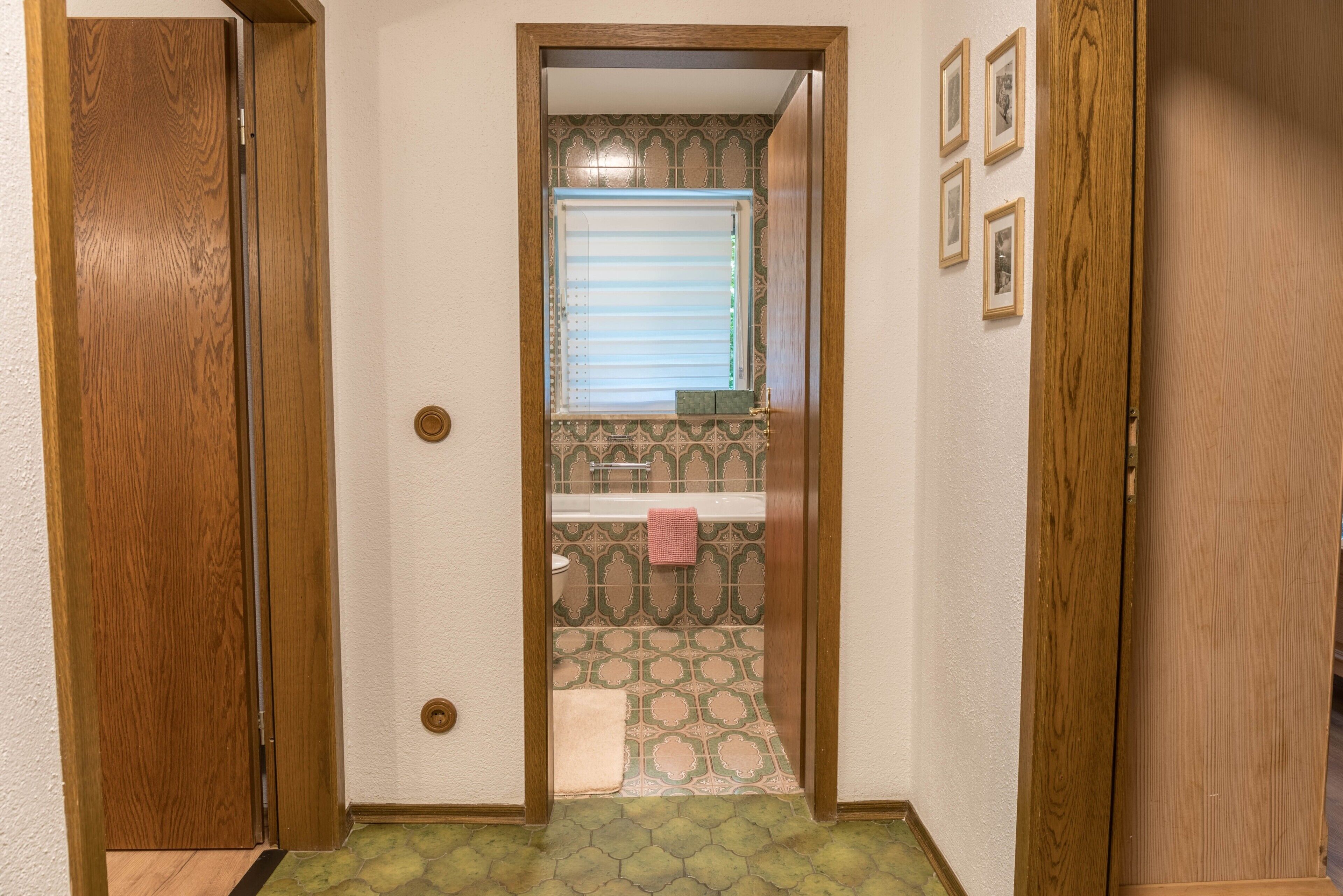 A hallway with a wooden door on the left, a window with blinds, and a patterned floor.