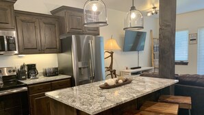 A modern kitchen with granite countertops, stainless steel appliances, wooden bar stools, and pendant lighting.