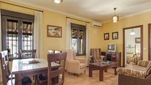Una sala de estar con una mesa de comedor, sillas, sofás, un televisor y un ventanal con cortinas.