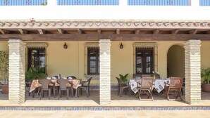 Un espacio comedor al aire libre techado, con mesas y sillas, piso de baldosas y barandilla.