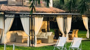 Un área de piscina con sillones reclinables, una zona de estar techada con cortinas y un edificio con tejado de azulejos.