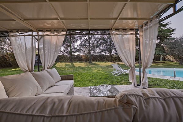 A covered outdoor seating area with a pool in the background.