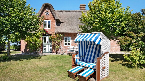 A traditional house with a thatched roof, a brick exterior, and a large window. In front, there is a striped beach chair and a small table.