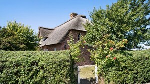 A thatched-roof house with a brick wall, surrounded by greenery and a white gate.