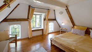 A bedroom with a sloped ceiling, wooden beams, a bed, a desk, and a window with curtains.