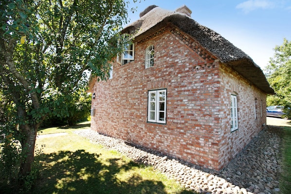 A brick house with a thatched roof, surrounded by trees and a gravel path.