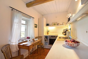 A kitchen with a wooden table, chairs, a bowl of fruit, a window with curtains, and a shelf with kitchen items.