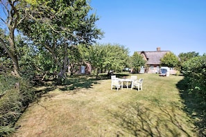 A grassy outdoor area with a table and chairs, surrounded by trees and a building with a thatched roof.