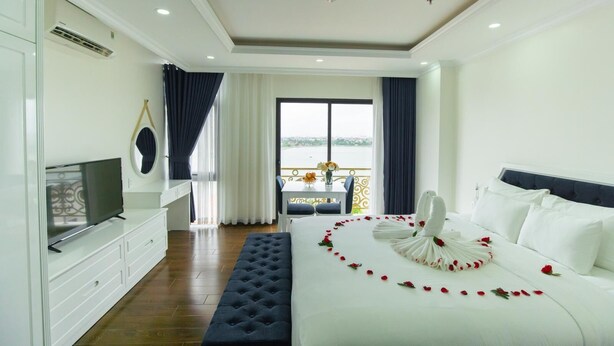 A hotel room with a bed, a TV, a balcony with a table and chairs, and a view of the water. - Queenie Hotel (Hai Phong)