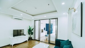 A modern living room with a white TV stand, a large plant, a teal sofa, and a glass door leading to a bedroom. - Queenie Hotel (Hai Phong)