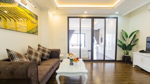 A living room with a brown sofa, white coffee table, and a large painting on the wall. There is a sliding glass door leading to a balcony with a view. - Queenie Hotel (Hai Phong)