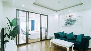 A modern hotel room with a wooden floor, a dark teal sofa, a white coffee table, and a large window with a view of the hotel lobby. - Queenie Hotel (Hai Phong)