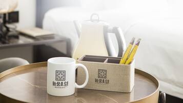 A neatly arranged hotel room with a bed, a bedside table with a lamp, a tray table with a mug, a box with pencils, and a pillow.