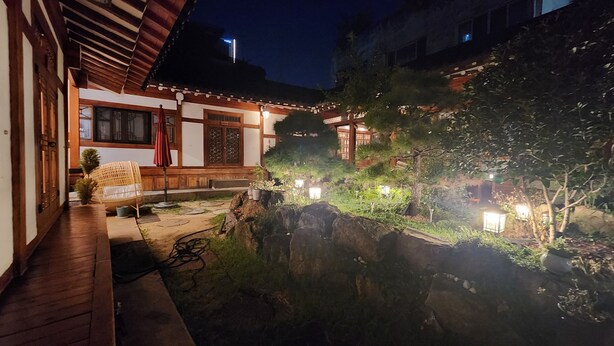 Un bâtiment de style japonais traditionnel, avec des poutres en bois et un toit recouvert de tuiles, un chemin de pierre et un petit jardin agrémenté de plantes et d’une lanterne.