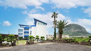 A modern hotel with a white and blue facade, surrounded by well-maintained gardens and a mountain backdrop. - Unicorn Pension (Seogwipo)