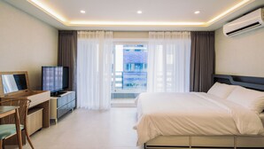 A hotel room with a bed, a desk with a TV, a window with curtains, and an air conditioning unit. - Unicorn Pension (Seogwipo)