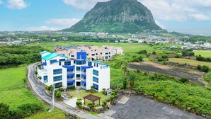 A modern building with multiple floors and balconies, surrounded by lush greenery and a prominent mountain in the background. - Unicorn Pension (Seogwipo)