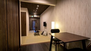 A room with a wooden floor, a dark wooden table, a black chair, a small table with a cushion, and a wall-mounted speaker. - Casa Grapp Tengachaya II (Osaka)
