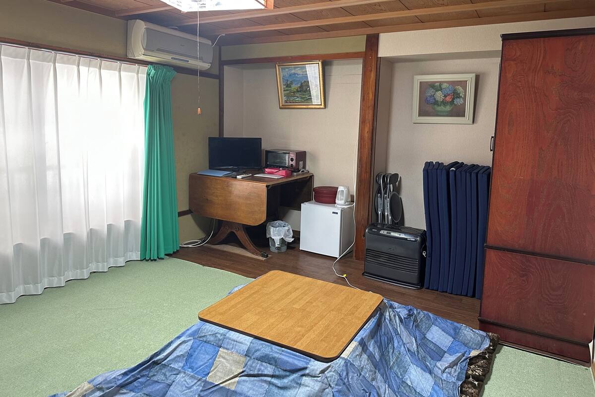 Een kamer met een bed, een bureau met computer, een kleine koelkast, een radiator en een raam met gordijnen.