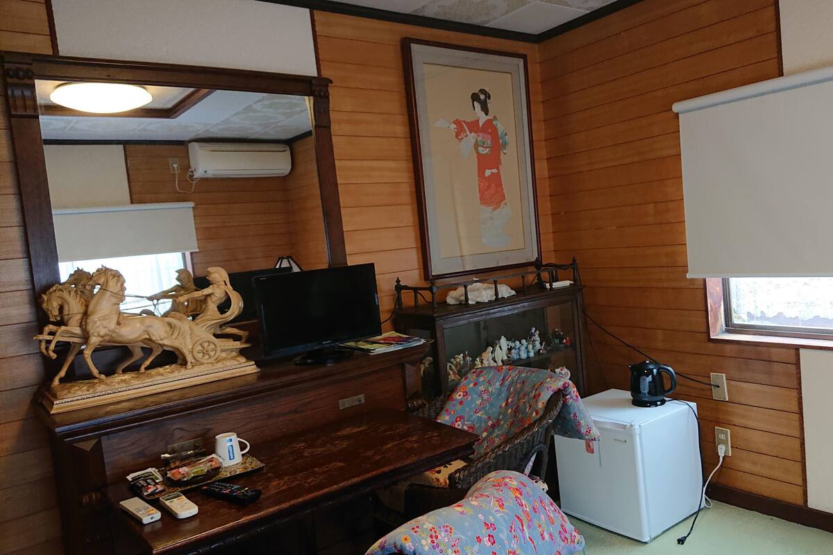 Een kamer met houten lambrisering, een televisie, een kleine koelkast, een houten kastje met beeldjes, een ingelijst beeld aan de muur en een raam met jaloezieën.