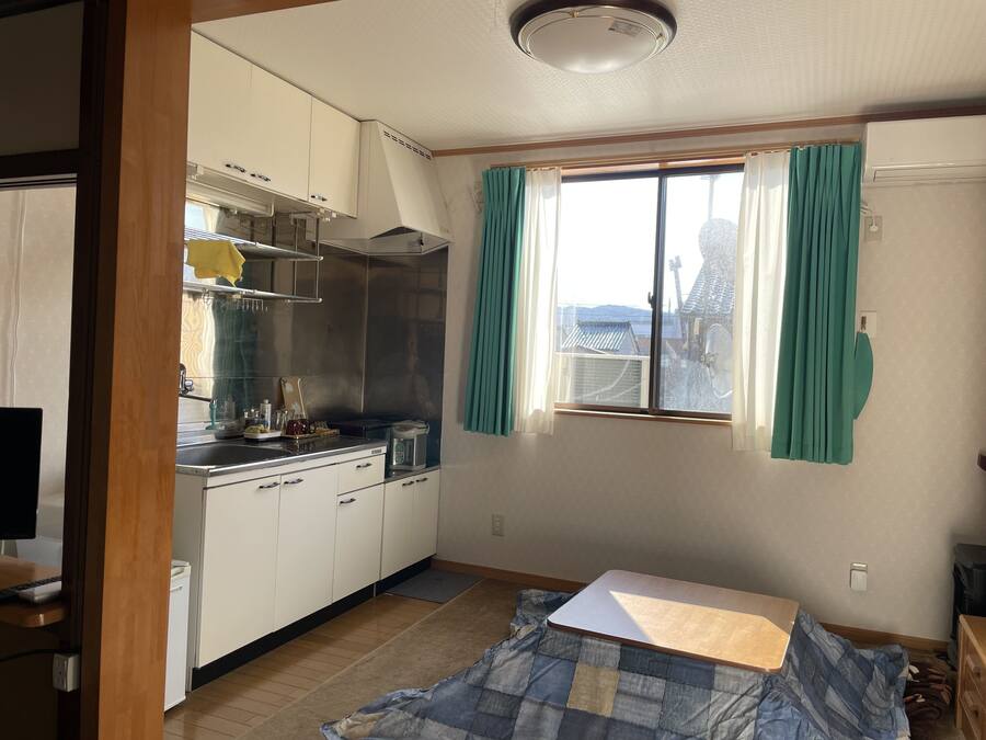 Une cuisine compacte avec des meubles blancs, une cuisinière et un évier. Il y a une fenêtre avec des rideaux turquoise et un lit recouvert d’une courtepointe bleue et beige.