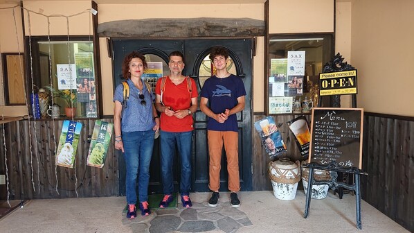 Three people standing in front of a building with a sign that says "Please come in!" and another sign indicating "Business hours 11:00 ~ 22:00".