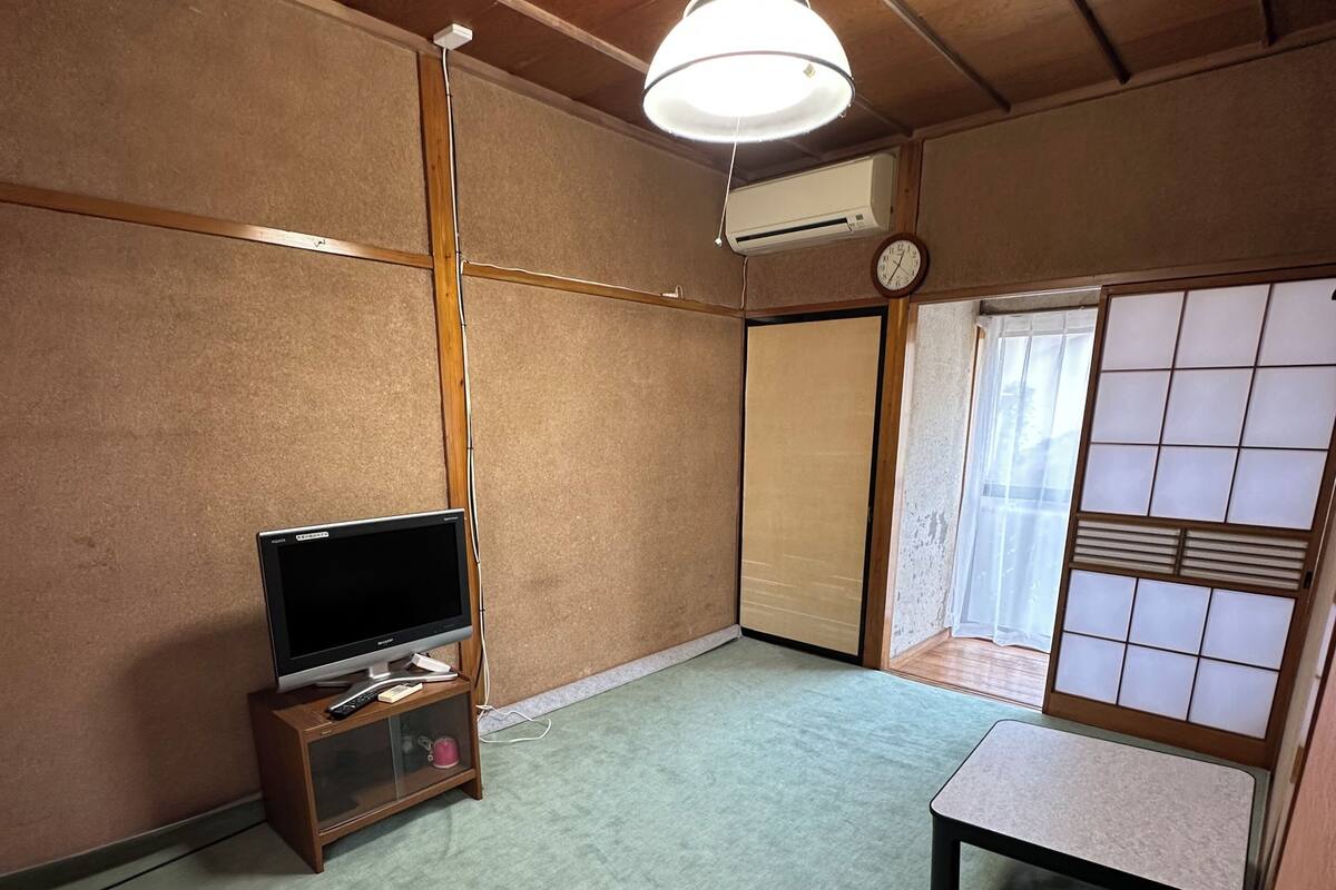 Een kamer met een televisie op een houten standaard, een klein tafeltje, een plafondventilator en een schuifdeur in shoji-stijl.