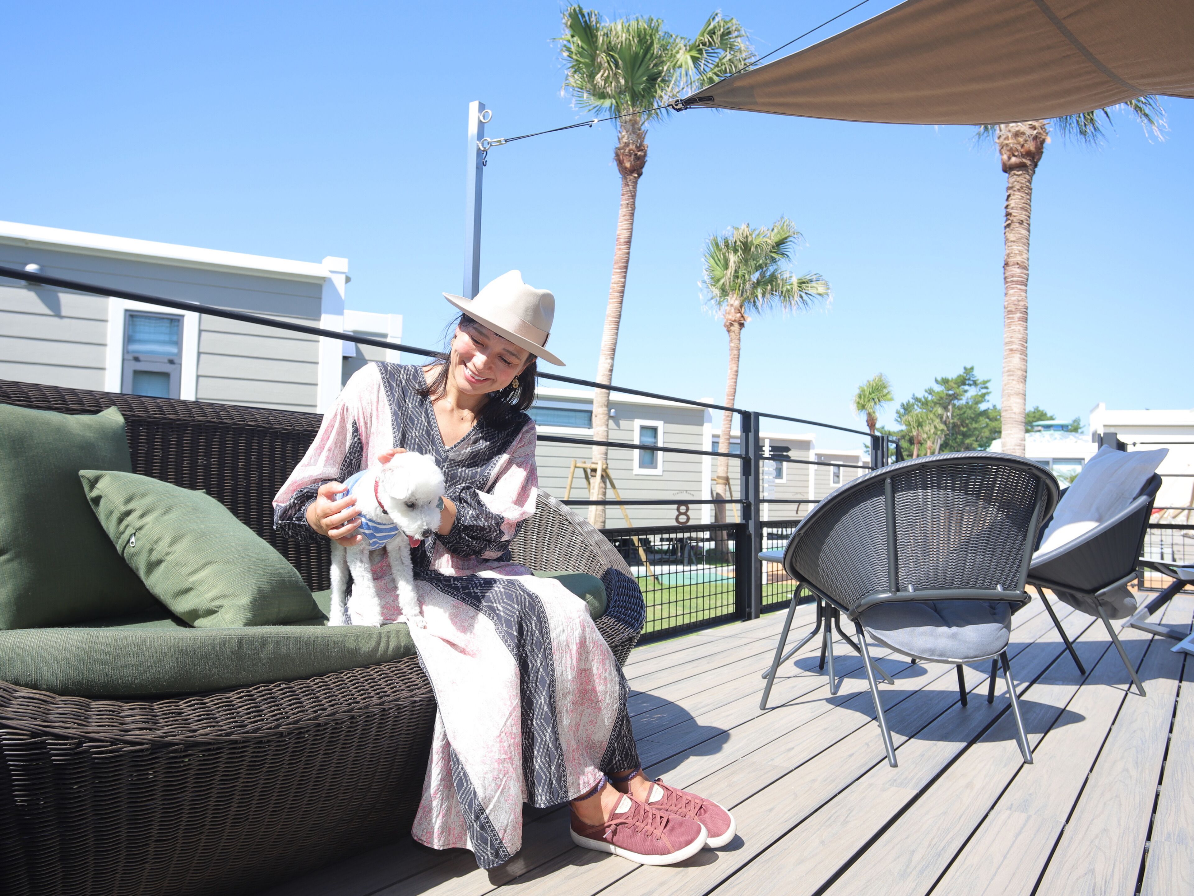 A person sitting on a wicker sofa with a dog, on a wooden deck with chairs and palm trees in the background.