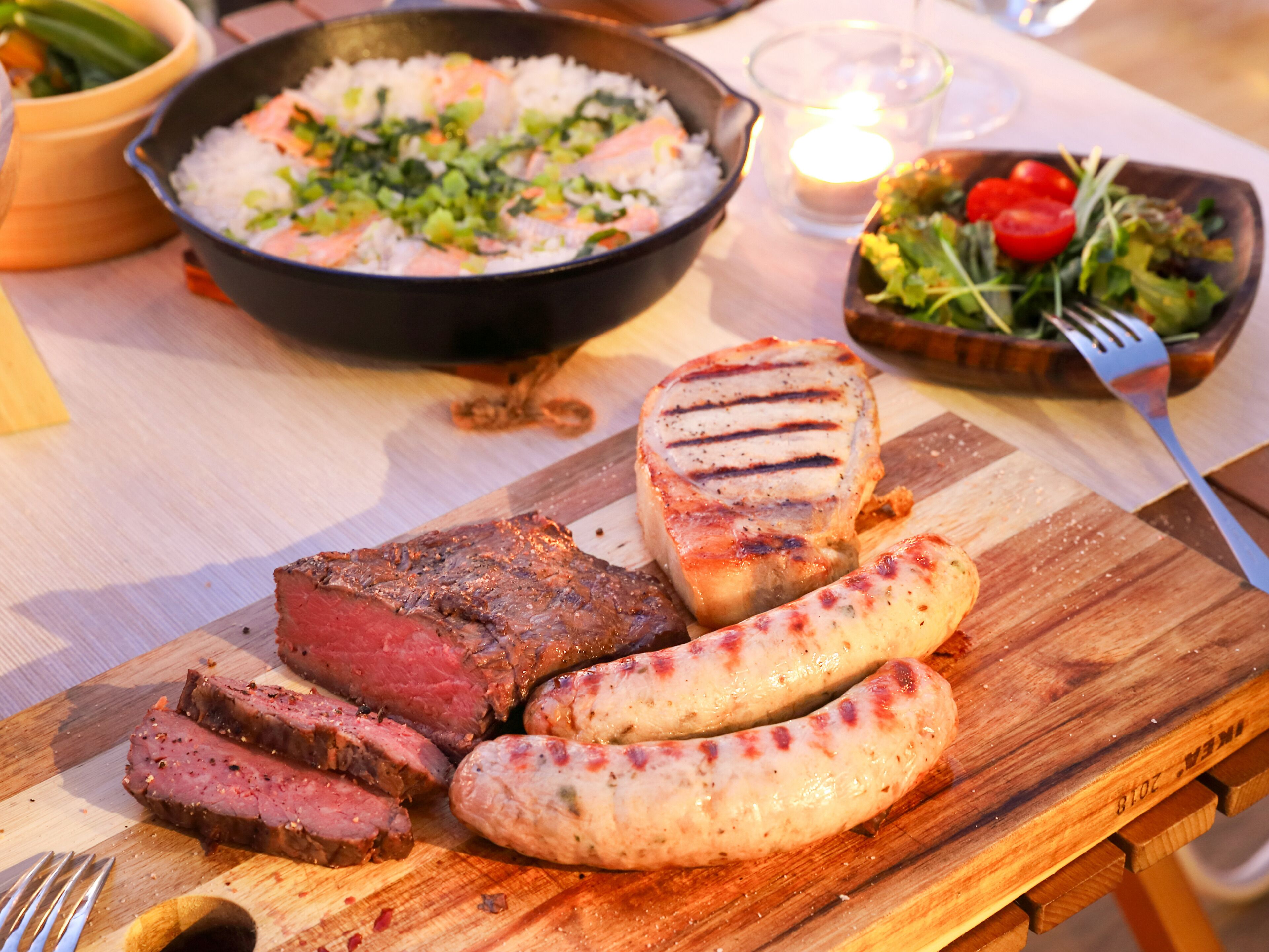 A wooden cutting board with sliced meat, sausages, and a grilled steak.