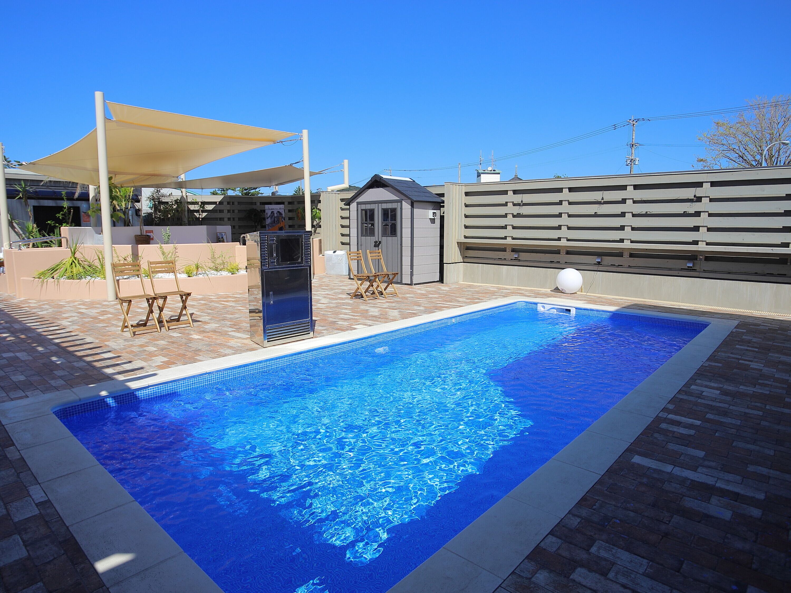 A rectangular swimming pool with a tiled edge, surrounded by a brick patio. There are two wooden chairs and a small table under a shaded area. A small shed is visible in the background.