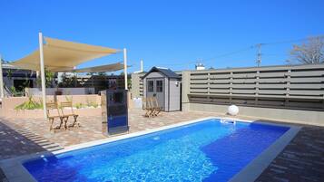 A rectangular swimming pool with a tiled edge, surrounded by a brick patio. There are two wooden chairs and a small table under a shaded area. A small shed is visible in the background.