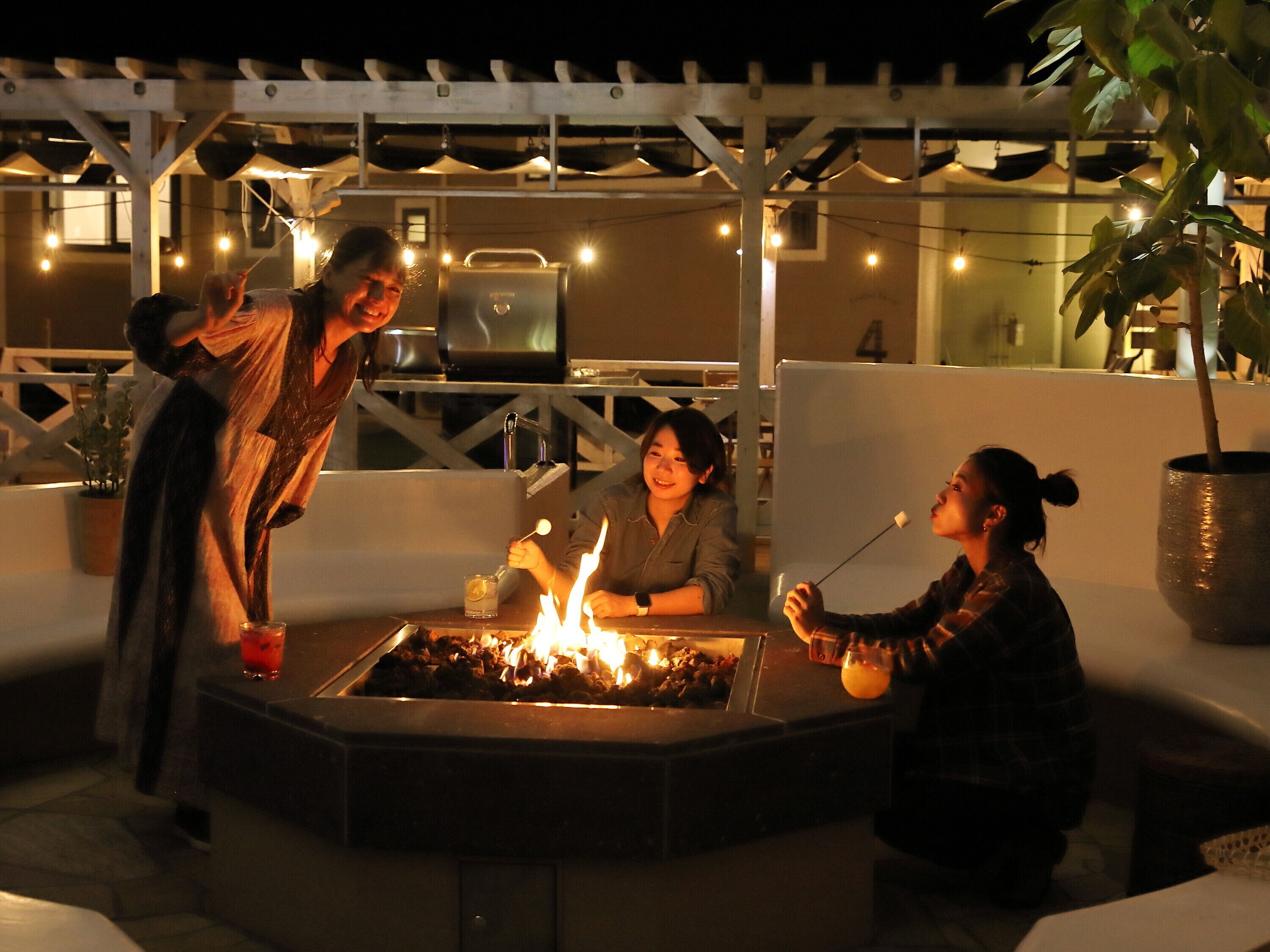 Un grupo de personas alrededor de un brasero en un entorno exterior con una pérgola arriba y luces colgantes.