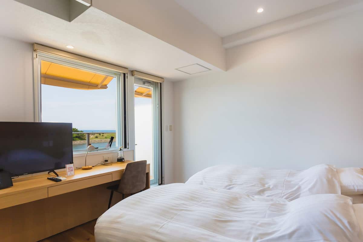 Une chambre avec un lit, un bureau avec un ordinateur et une télévision. Il y a une fenêtre donnant sur l’extérieur.